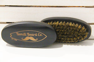 Image of two Bondi Beard Co. Boar Bristle Brushes, showing both sides, one with branding, the other with coarse boar bristles. Sitting on white wooden surface.