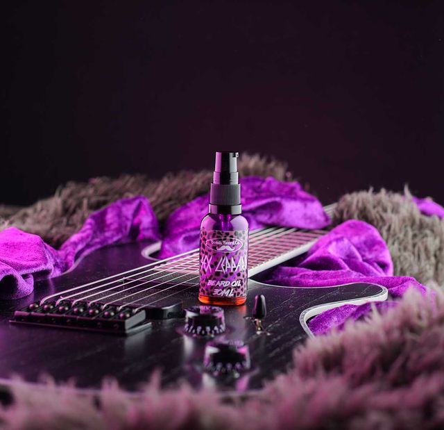 Image of Bondi Beard Co. Sex Leppard beard oil with bright purple leopard print label, spray bottle is placed on a black electric guitar and bright purple velvet and faux fur fabrics in background. 